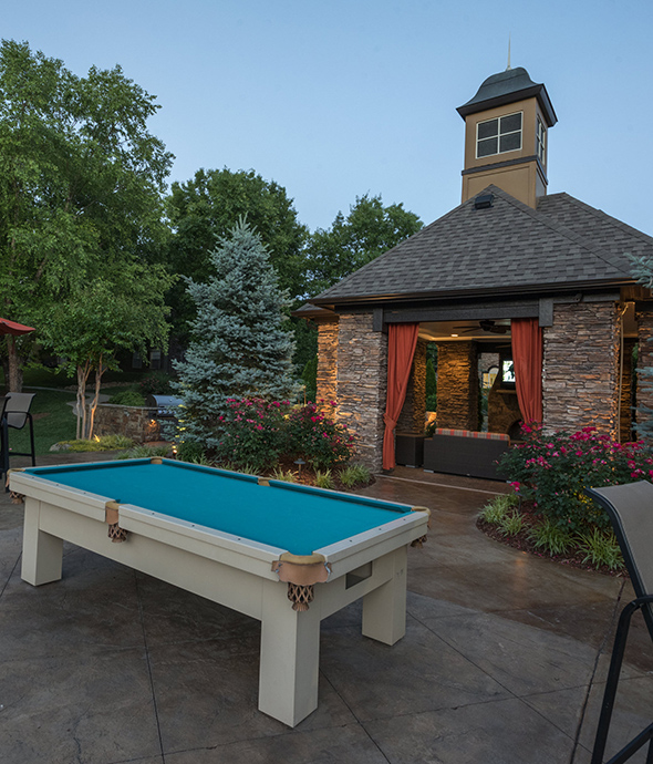 Outdoor area with stone pavers, large brick gazebo, outdoor pool table, and lush landscaping with tall trees.