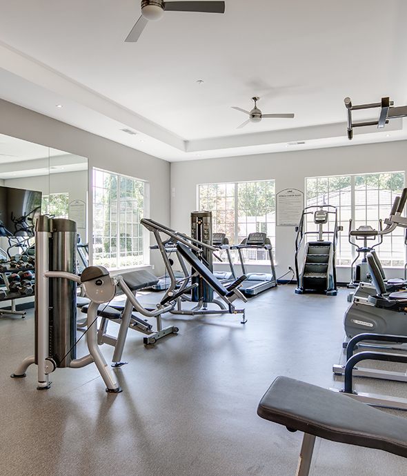 Fitness center with mirror walls, cardio and weight equipment, and large windows with natural light.